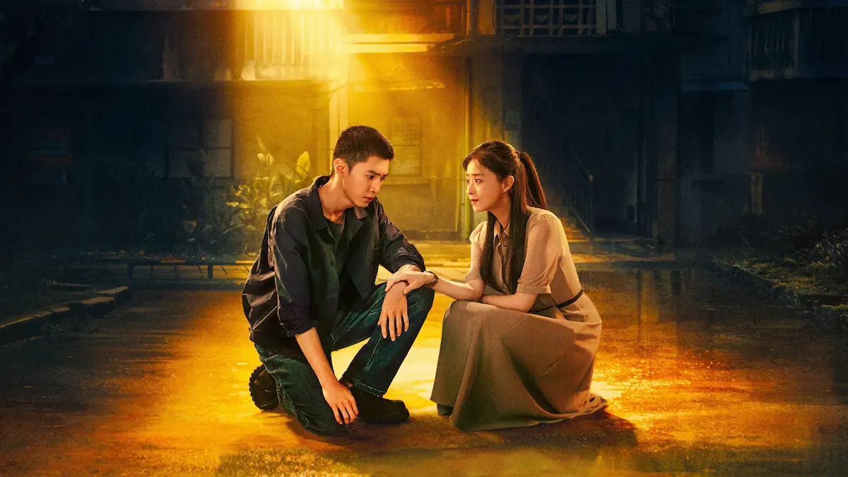 A young man crouching on a wet alley street looking down while a woman in a beige dress kneels beside him, holding his arm under warm yellow streetlight