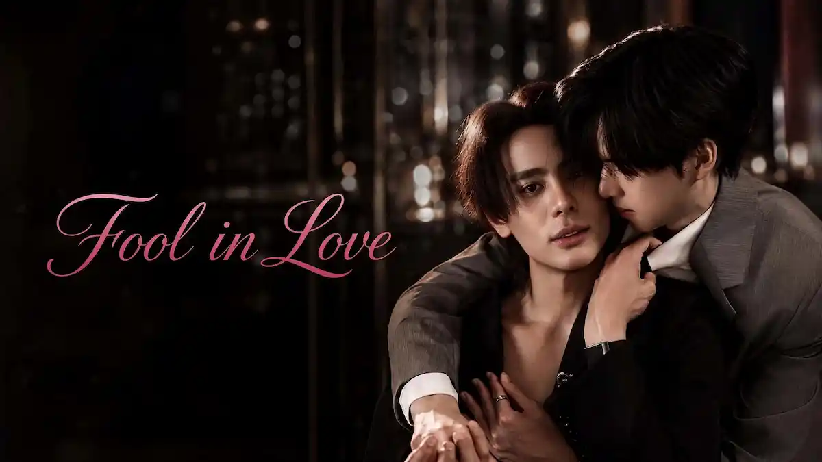 A man in a dark jacket held intimately from behind by another man in a grey suit, with cursive title text reading Fool in Love